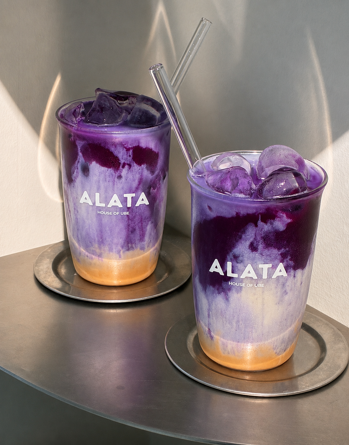 Two tall clear cups filled with purple and lavender ALATA dirty iced ube latte drinks and ice, each with a clear straw, resting on round metal coasters on a gray tabletop. The cups are branded ALATA.
