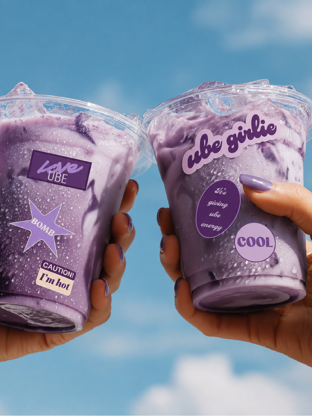 Two hands holding clear plastic cups filled with a purple ube iced latte drink against a blue sky background. The cups have purple labels and graphics, including one that says 'cube girlie' and another that says 'BOMB CAUTION! I'm hot'.