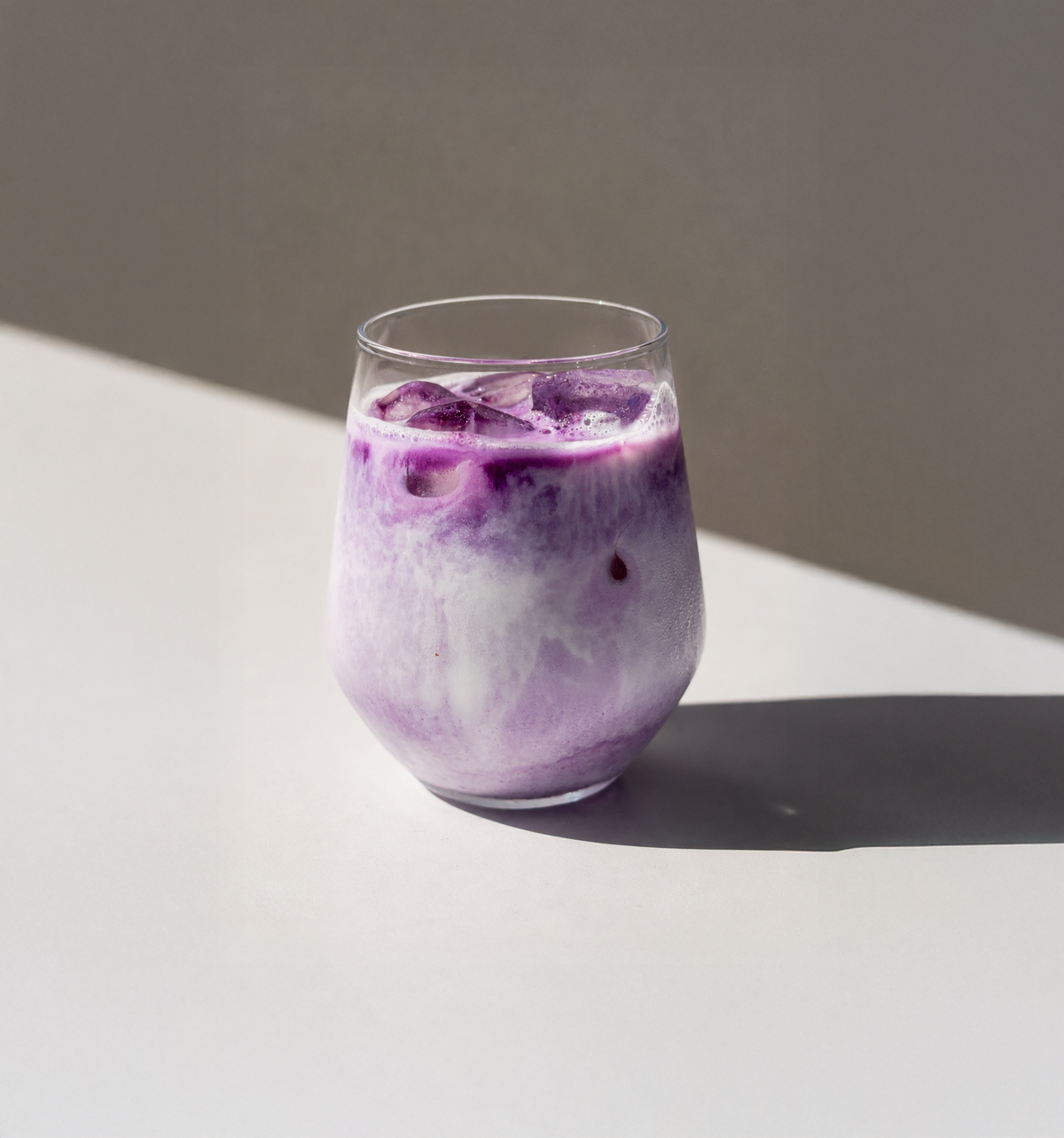 A clear stemless glass filled with a purple-and-white iced ALATA ube latte drink and large ice cubes, placed on a white surface with a soft shadow against a warm gray background.