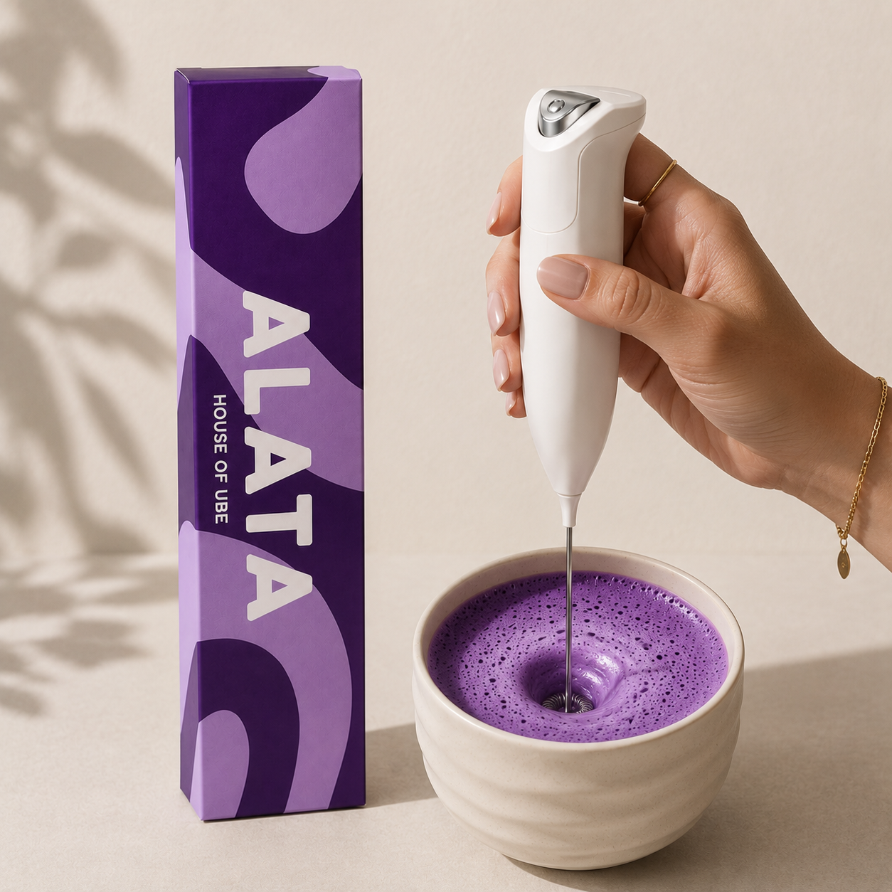 A hand holds a white handheld milk frother over a white bowl filled with purple frothed milk, next to a purple ALATA product box on a warm beige surface.